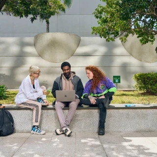 Three people sit outdoors on a campus bench, collaborating while viewing a laptop.